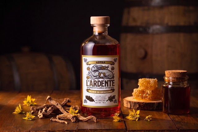 Bottiglia di Astra Elisir di Genziana de L'Ardente Micro Distilleria Artigiana su banco in legno antico, con radici di genziana, favo di miele e fiori gialli